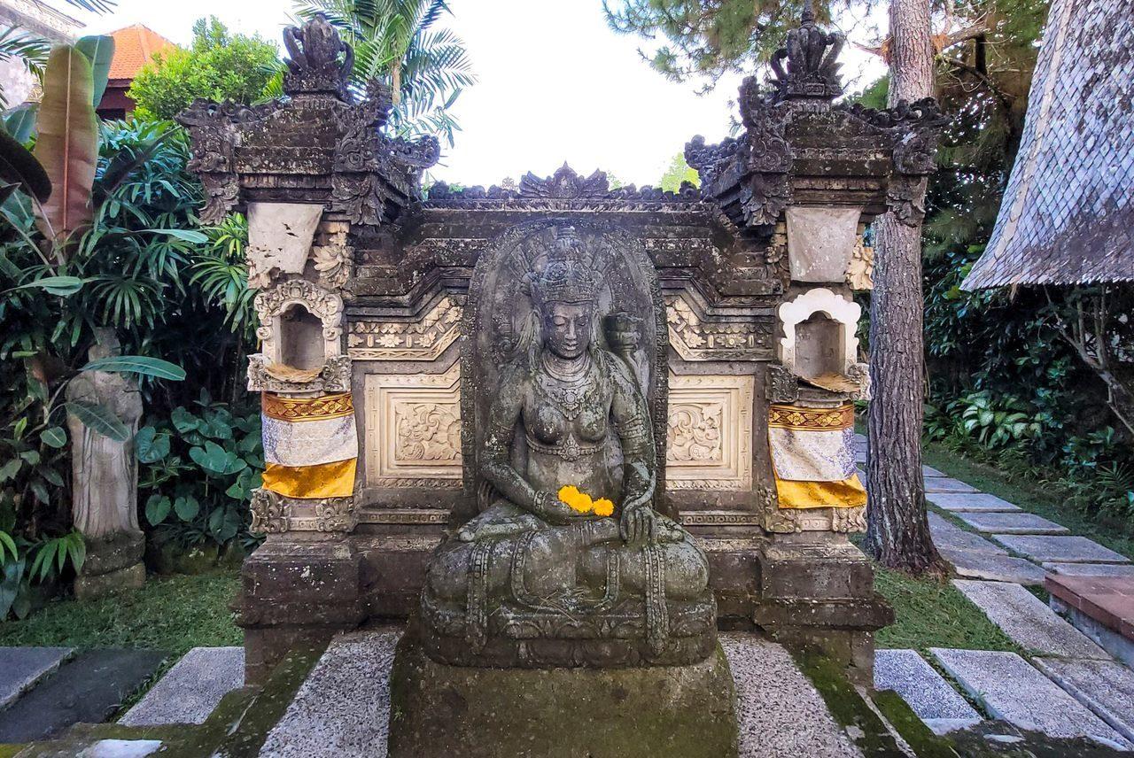 A carved stone aling-aling wall in a Balinese garden, with a seated figure in a niche and offerings at its foot