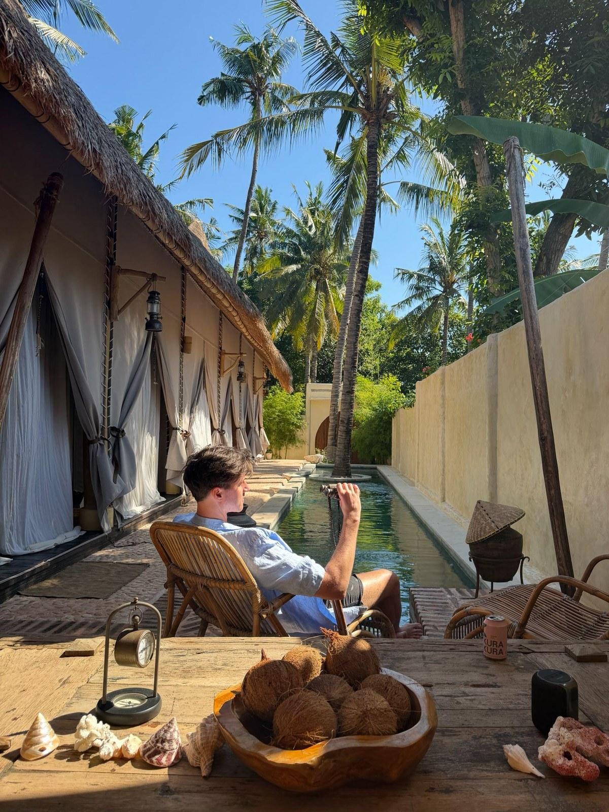 Danny in a rattan chair beside a long pool under coconut palms, a bowl of coconuts on the table
