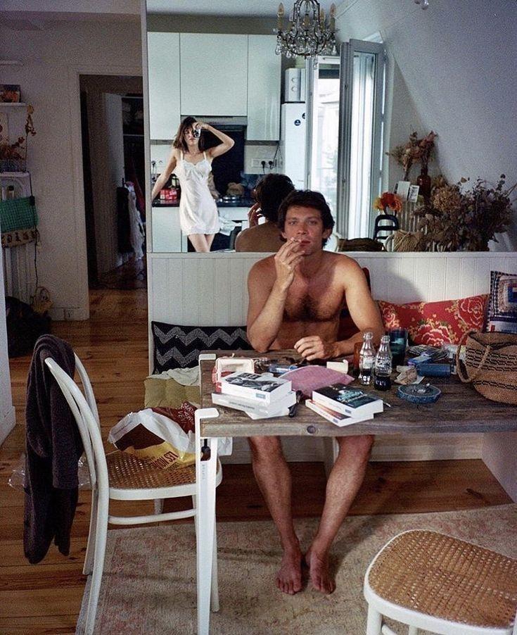 A man in his underwear smoking at a kitchen table, a woman posing in the background