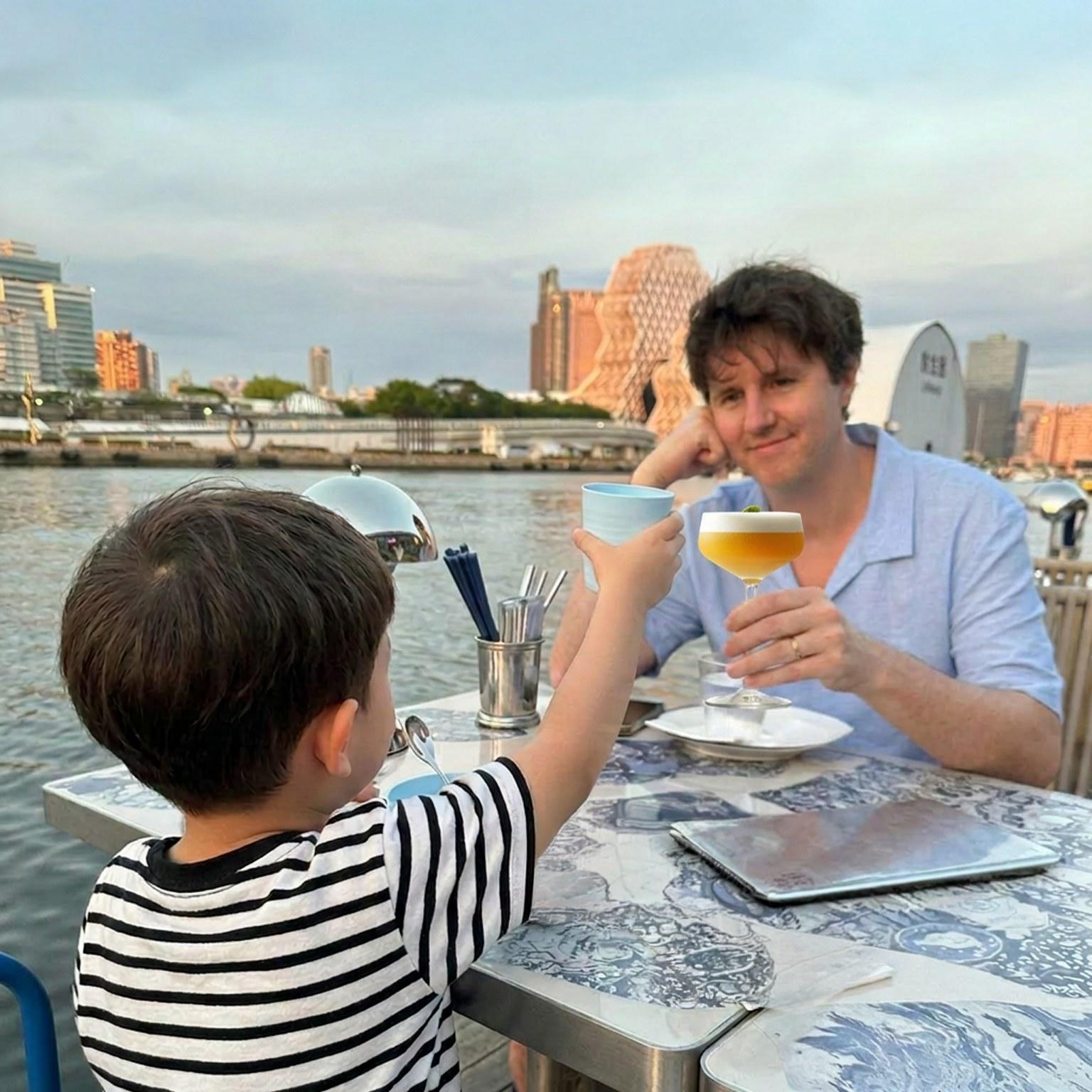 Danny and his son raising glasses at a waterside table in Kaohsiung at dusk